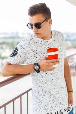 Klasik güneş gözlüklü havalı hippi beyaz tişörtlü adam kahve tutuyor ve balkonda duruyor.