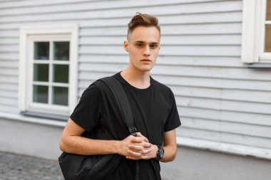 Moda siyah tişörtlü yakışıklı genç adam. Sırt çantası eski ahşap bir binanın yanında.