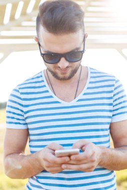 Çizgili tişörtlü, saç stili ve sakallı yakışıklı hippi plajda akıllı telefon kullanıyor.