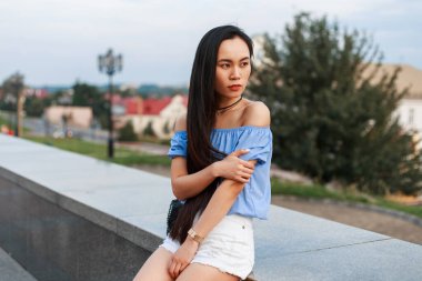 Güzel, modaya uygun, uzun saçlı, yazlık elbiseli ve şortlu Asyalı bir kız şehirde oturuyor.