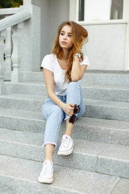 Şık, kızıl saçlı, modaya uygun giyinen, tişörtlü ve kot pantolonlu güzel bir kadın model şehirde merdivenlerde oturuyor.