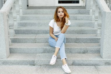 Şık, kızıl saçlı, modaya uygun beyaz tişörtlü, kot ve spor ayakkabılı bir kız model şehrin basamaklarında oturuyor.