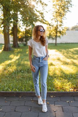 Kırmızı saçlı, güneş gözlüklü, tişörtlü ve kot pantolonlu güzel bir kadın parkta poz veriyor.