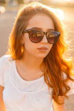 Şık beyaz tişörtlü, kırmızı saçlı, moda güneş gözlüklü şık bir kadın model gün batımında şehirde yürüyor.