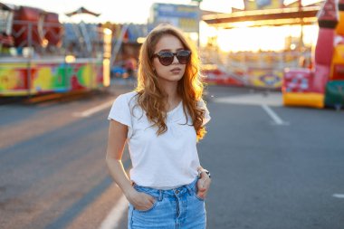 Güneş gözlüklü, beyaz tişörtlü ve kot pantolonlu güzel bir manken şehirde gün batımında yürüyor.