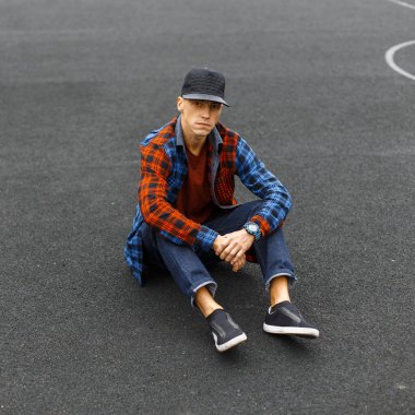 Havalı yakışıklı moda öğrencisi siyah şapkalı, gömlekli ve kot pantolonlu asfaltta oturuyor.