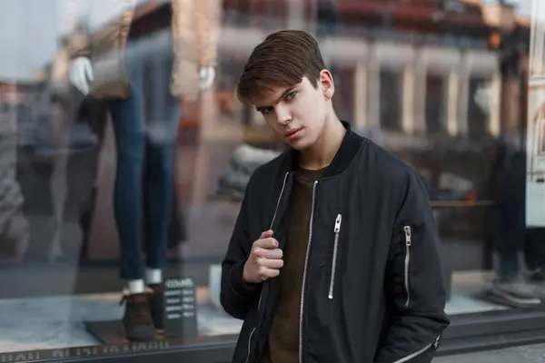 stock image Handsome fashion guy with a hairstyle in a black stylish jacket stands near a store window outdoors