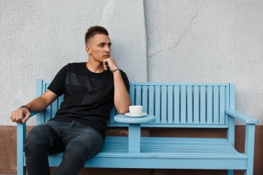 Siyah elbiseli yakışıklı bir adam bankta oturur ve kahve içer.