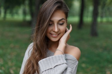 Moda örgü süveter giymiş sevimli bir gülümseme mankeni olan genç ve mutlu bir kız doğada parkta yürüyor.