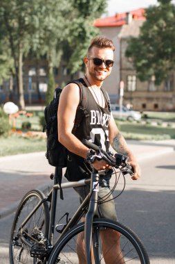 Güneş gözlüklü yakışıklı hippi bir bisikletin yanında duruyor ve güneşli bir günde şehirde eğleniyor.