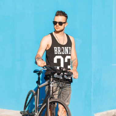 Moda gözlüklü, yaz kıyafetleriyle sokakta mavi arka planda bisikleti olan yakışıklı, hippi bir bisikletçi.