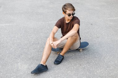 Moda gözlüklü havalı genç hippi yazın giydiği giysiler sokakta asfaltta kaykayın üzerinde oturuyor.