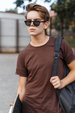 Güneş gözlüklü, şık tişörtlü, sırt çantalı ve sokakta kaykaylı genç, güzel bir moda adamı.