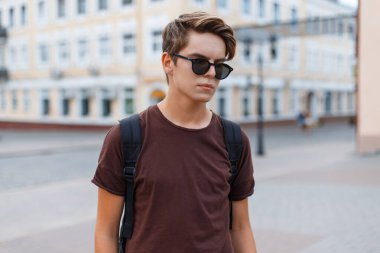 Şık, yakışıklı, saç stili, güneş gözlüklü, tişörtlü ve sırtında sırt çantası olan genç bir adam şehirde dolaşıyor.