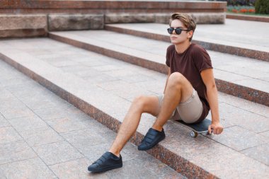 Moda tişörtlü, güneş gözlüklü, şık, yakışıklı genç adam şehirde bir kaykayın üzerinde oturuyor.
