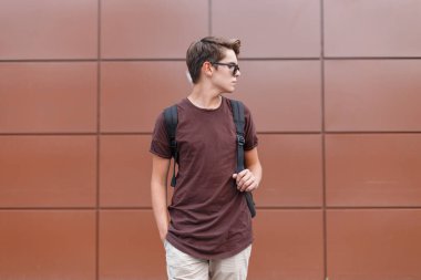 Moda tişörtlü, kahverengi arka planda sırt çantası olan şık güneş gözlüklü yakışıklı bir genç.