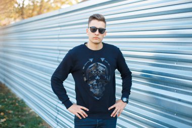 Moda gözlüklü, yakışıklı bir genç adam. Siyah kazaklı. Sokakta metal arka planda.