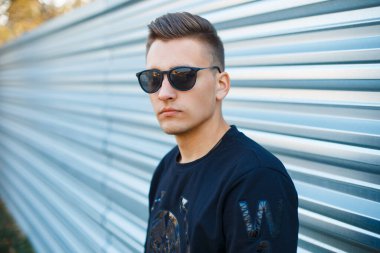 Modaya uygun güneş gözlüklü yakışıklı bir adam. Metal bir arka planda siyah bir kazakla caddede.