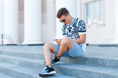 Yazlık giysiler içinde yakışıklı genç adam şehirde oturmuş müzik dinliyor.