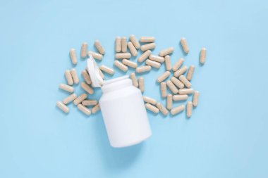 Açık mavi arka planda bej kapsülleri dökülmüş beyaz plastik şişe. İlaç, sağlık, takviye veya vitamin kavramı. Tıbbi temalar için temiz, minimalist kompozisyon.