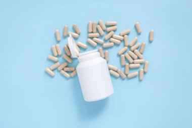 Pastel mavi arka plana saçılmış doğal destek kapsülleri olan beyaz bir şişe. Fotokopi alanı ile sağlık, sağlık ve beslenme kavramı.