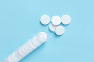 Mavi kapaklı şeffaf plastik bir tüpten yayılmış beyaz yuvarlak tabletler. Temiz mavi arka planda sağlık ve ilaç konsepti