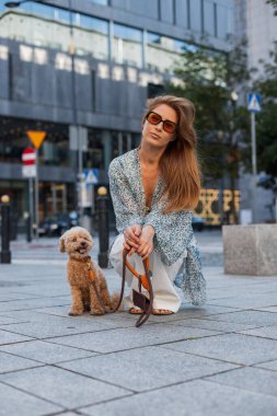 Şık yaz kıyafetli bir kadın şehirde köpeğini tasmayla gezdiriyor. Rahat bir ortam, modern şehir arkaplanı ve ev sahibiyle evcil hayvan arasında bir bağlantı anı..