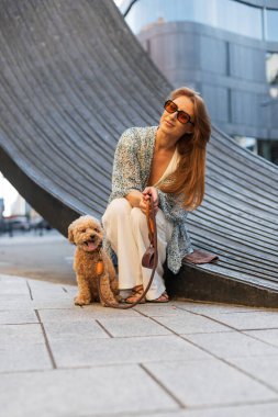 Genç bir kadın modern şehir ortamında zıplayan kanişiyle eğleniyor. Evcil hayvanla bağ kurma anı. Neşeli yürüyüş, yaz havası ve arkadaşlık.