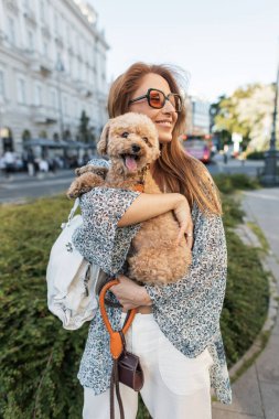 Şehrin göbeğinde köpeğini kucaklayan şık bir kadın. Kaniş rahat ve mutlu görünüyor. Sahibi ve evcil hayvanı arasındaki sevgi bağını yakalıyor..