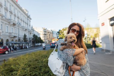 Genç bir kadın şehirde yürürken tüylü kanişini kucaklıyor. Aydınlık giyinmiş, yaz kıyafetleri içinde, aydınlık bir günde mutluluk ve şefkat yayıyor.