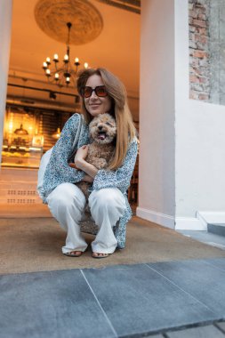 Yazlık kıyafetli genç bir kadın küçük köpeğiyle şık bir elbiseden çıkıyor. Şehir hayatı, evcil hayvan arkadaşlığı ve bu samimi şehir anında sıradan moda karışımı. Kızıl saçlı güzel kadın.