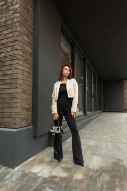 Modern bir binanın yanında duran bej ceketli ve siyah kıyafetli şık genç bir kadın. Kendine güvenen profesyonel görünüş.
