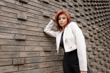 Dışarıda şık bir kadın, siyah bir elbise ve beyaz bir ceket giyiyor. Saçlarında rüzgar, gelişigüzel şehir ortamı, modern tuğla arka plan. Ceketli, şık, kızıl saçlı, güzel bir kadın.