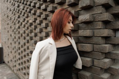 Modern genç kadın geometrik tuğla duvarın yanında duruyor, şık bir elbise giymiş. Şık bir sokak modası. Tuğla duvarın yanında ceketi olan şık, kızıl saçlı güzel bir kadın..