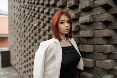 Modern genç kadın geometrik tuğla duvarın yanında duruyor, şık bir elbise giymiş. Şık bir sokak modası. Tuğla duvarın yanında ceketi olan şık, kızıl saçlı güzel bir kadın..