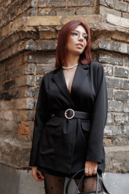 Tuğlalı bir duvarın önünde şık siyah bir elbise giymiş kendine güvenen güzel bir kadın. Moda, entellektüellik ve sokak stili..