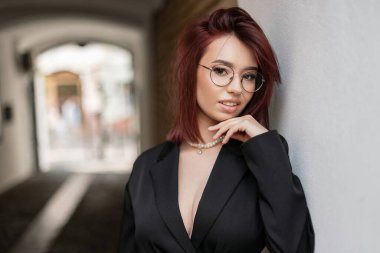 Kısa kızıl saçlı, gözlüklü, siyah ceketli dar bir sokakta duvara yaslanmış çekici bir genç kadın. Şehir modası ve güzellik kavramı.