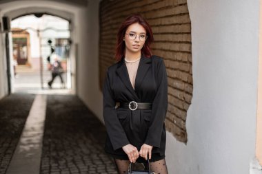 V yaka ceketli ve inci kolyeli şık bir kızıl, şehrin kemer yolundaki beyaz bir duvara yaslanıyor. Sofistike sokak tarzı bir portre..