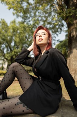 Siyah elbiseli ve desenli taytlı zarif bir kadın güneşli bir parkta güven içinde poz veriyor. Modern moda ve doğal ışık ile açık hava yaşam tarzı.