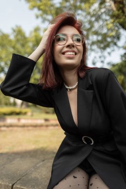 Şehir parkında güneşin tadını çıkaran zarif siyah giyimli, inci kemer ve kolyeli genç bir model. Doğal ortamda modaya uygun bir görünüm. Mutlu gülümseyen kız