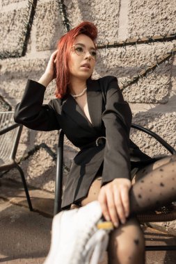 Kırmızı saçlı, güneş gözlüklü, şık siyah elbiseli şık bir kadın model dışarıda duvarın yanında oturmuş, güneşin tadını çıkarıyor ve gösterişli bir moda ifadesi sergiliyor..