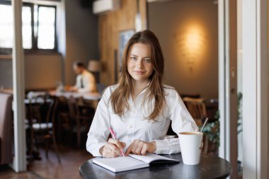 Beyaz gömlekli genç bir kadın kahve ve telefonuyla bir kafede notlar yazıyor. Konforlu çalışma alanı atmosferi, çalışma, planlama, günlük tutma veya üretkenlik kavramları için ideal.