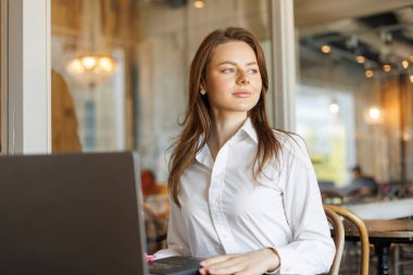 Kafe masasında dizüstü bilgisayar kullanan kendine güvenen bir kadın. Klasik kıyafet, dijital aletler ve kahve modern profesyonellerin çalışma alanı ve esnek çalışma rutinini gösteriyor.