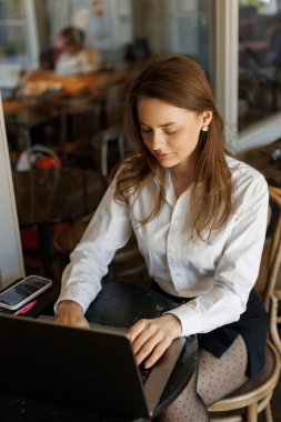 Beyaz gömlekli bir iş kadını şık bir kafede dizüstü bilgisayar kullanıyor. Başarı, bağımsızlık ve dijital iş yaşam tarzının sembolü.