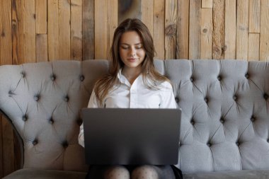 Şık bir koltukta otururken dizüstü bilgisayarda çalışan genç bir kadın. Modern ahşap iç mekan, uzaktan çalışma, serbest yaşam tarzı, sakin ve üretken atmosfer.