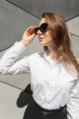 Güneş gözlüğü, beyaz bluz ve siyah etek giyen zarif bir kadın model gün ışığında geometrik gri bir duvarın önünde dikiliyor. İş kadını. Güzel bayan.