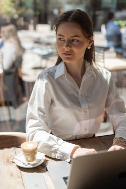 Kafe masasında dizüstü bilgisayarda çalışan genç bir iş kadını kahve ve akıllı telefonuyla. Gün ışığı çalışma alanı, uzak iş konsepti, gündelik modern yaşam tarzı.