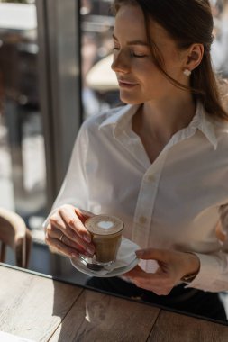 Kahve molası veren, şık giyinmiş bir kadın. Beyaz gömlekli genç bir kadın bir kafede kahve içiyor. Kahve içen genç iş kadını.