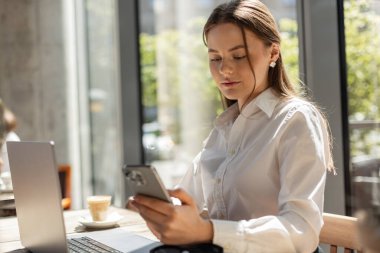 Beyaz gömlekli kadın modern ofiste akıllı telefon ve dizüstü bilgisayarla birden fazla iş yapıyor. Dijital çalışma, iletişim ve uzaktan üretkenlik kavramı. Ofis hayatı: telefonu kontrol eden kadın