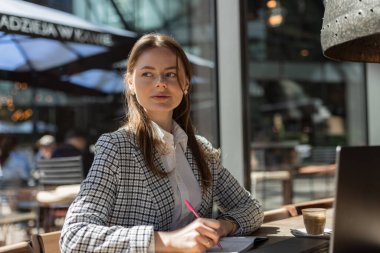 Serbest çalışan ya da öğrenci, güneşli bir kahve dükkanında dizüstü bilgisayarın yanında not alıyor. Masada kaktüs bitkisi ve kapuçinoyla çalışan şık bir kadın.